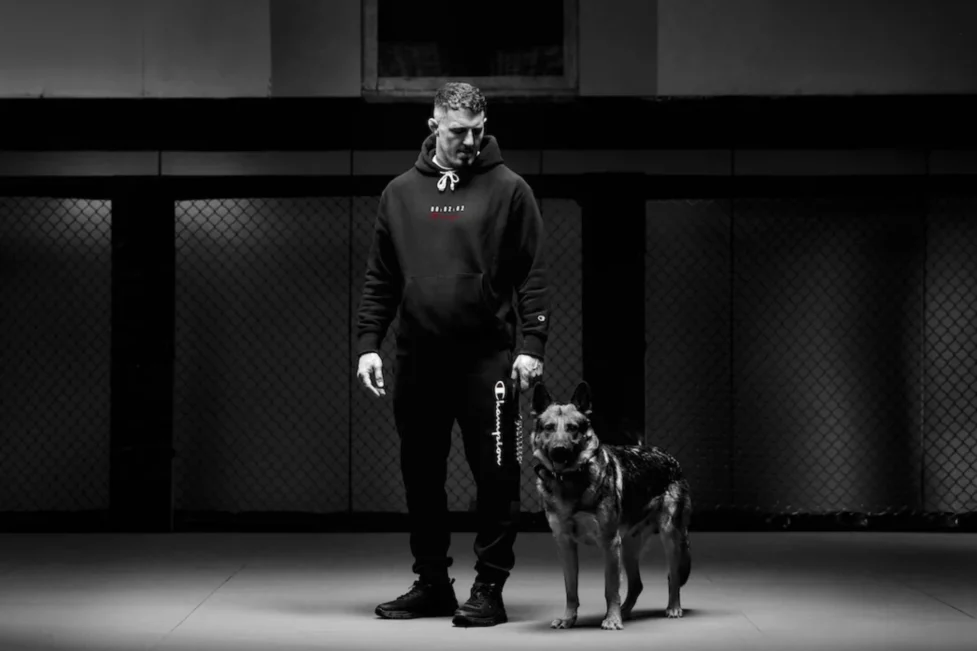 Retrato cinematográfico en blanco y negro de Tom Aspinall, campeón de la UFC, vistiendo una hoodie clásica de Champion junto a un perro pastor en un entorno de entrenamiento.