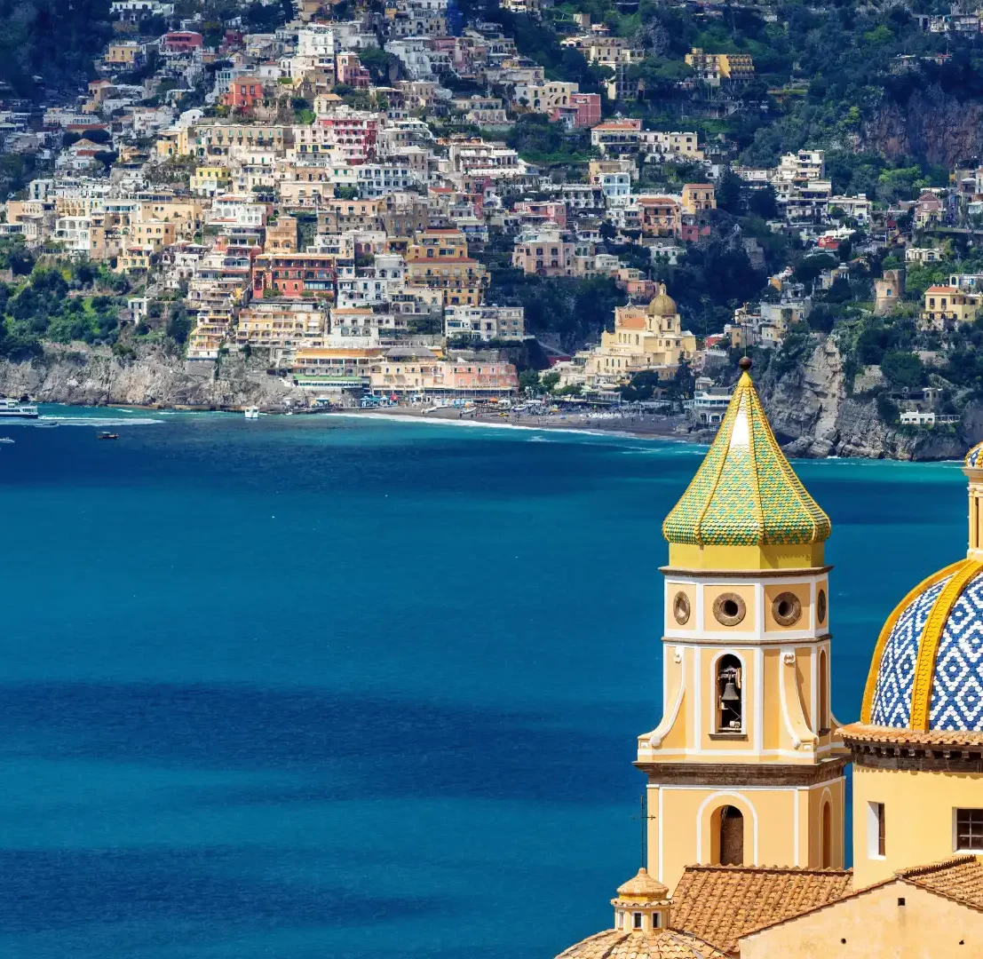 Vista panorámica de la cúpula de mayólica amarilla y azul de la Iglesia de la Anunciación en Salerno, Italia, con el mar Mediterráneo y la Costa Amalfitana de fondo.