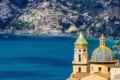 Vista panorámica de la cúpula de mayólica amarilla y azul de la Iglesia de la Anunciación en Salerno, Italia, con el mar Mediterráneo y la Costa Amalfitana de fondo.