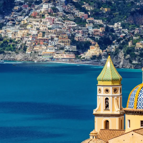 Vista panorámica de la cúpula de mayólica amarilla y azul de la Iglesia de la Anunciación en Salerno, Italia, con el mar Mediterráneo y la Costa Amalfitana de fondo.