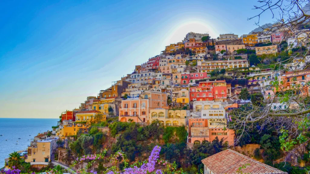 Para esa imagen icónica de Positano, donde las casas de colores parecen desafiar la gravedad incrustadas en el acantilado, el texto alternativo debe evocar esa sensación de drama arquitectónico y lujo mediterráneo que define a Cultura Foodie.

Aquí tienes tres opciones de texto alt, desde la más descriptiva hasta la más evocadora para el SEO de lujo:

Opción 1: Descriptiva y Técnica (Recomendada para SEO)
Casas de colores pastel incrustadas verticalmente en el acantilado de piedra caliza de Positano, Costa Amalfitana, vistas desde el mar Tirreno en un día soleado.