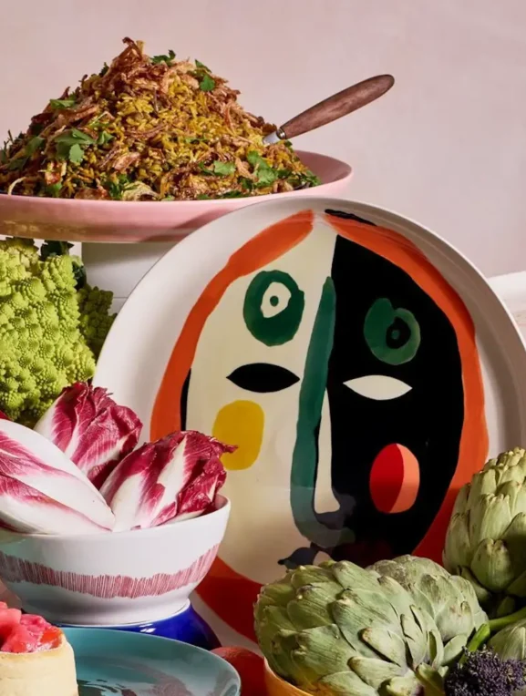 Mesa servida con la colección de vajilla colorida diseñada por Yotam Ottolenghi, platos en tonos azul, amarillo y rosa con platillos gourmet servidos.