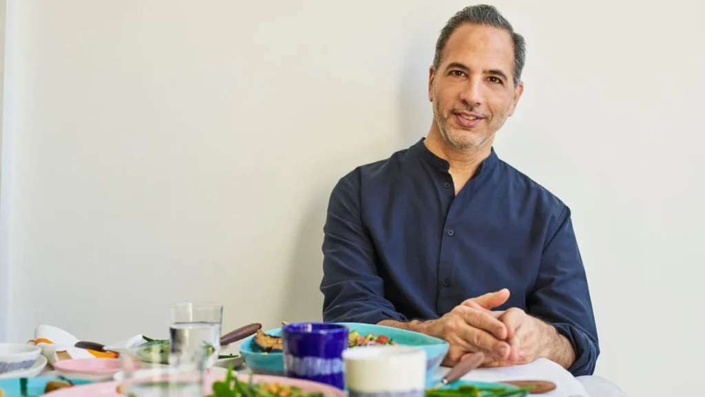 El chef Yotam Ottolenghi posando junto a una mesa llena de platillos gourmet servidos en su propia línea de vajilla de colores vibrantes.