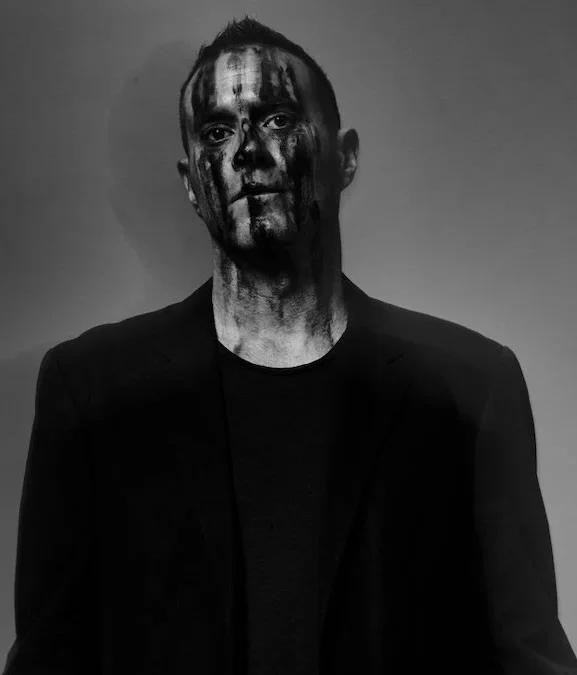 James Lavelle, fundador del colectivo UNKLE, con el rostro intervenido con pintura artística de estilo geométrico abstracto, iluminado por luces de neón en un entorno de estudio.