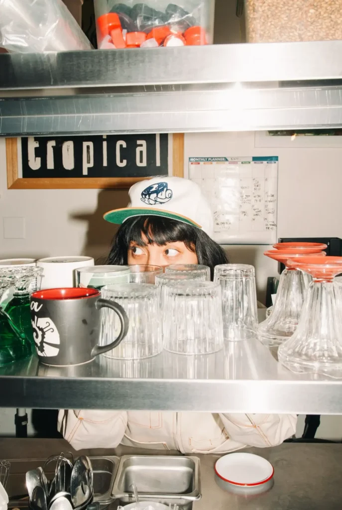 Barista detrás de una barra vistiendo la gorra para baristas de la nueva colección de moda Oatly x Kids of Inmigrants.