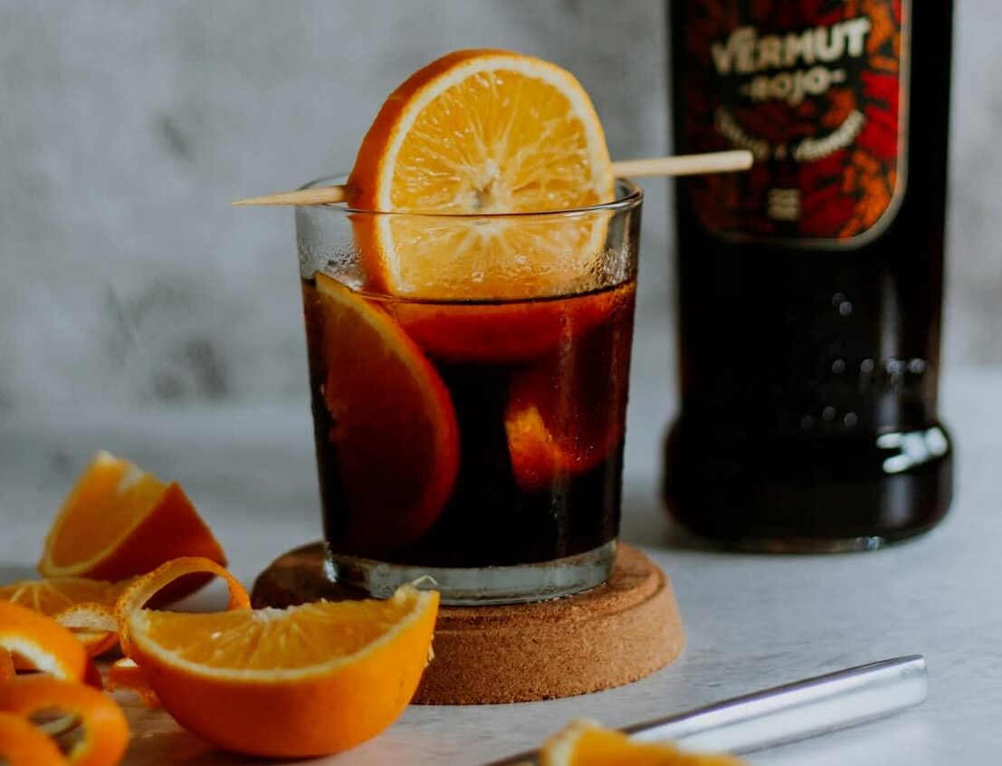 Copa de vermut rojo con hielo y rodaja de naranja fresca junto a botella de cristal en primer plano para el aperitivo.
