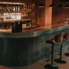 Barra curva de azulejos oscuros con bancos altos y botellas de vino en Espacio Mariatinto, Valle de Guadalupe, Baja California.