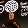 Presentadora vestida de negro frente al logotipo oficial de The World's 50 Best Bars 2026 durante la ceremonia de premiación.